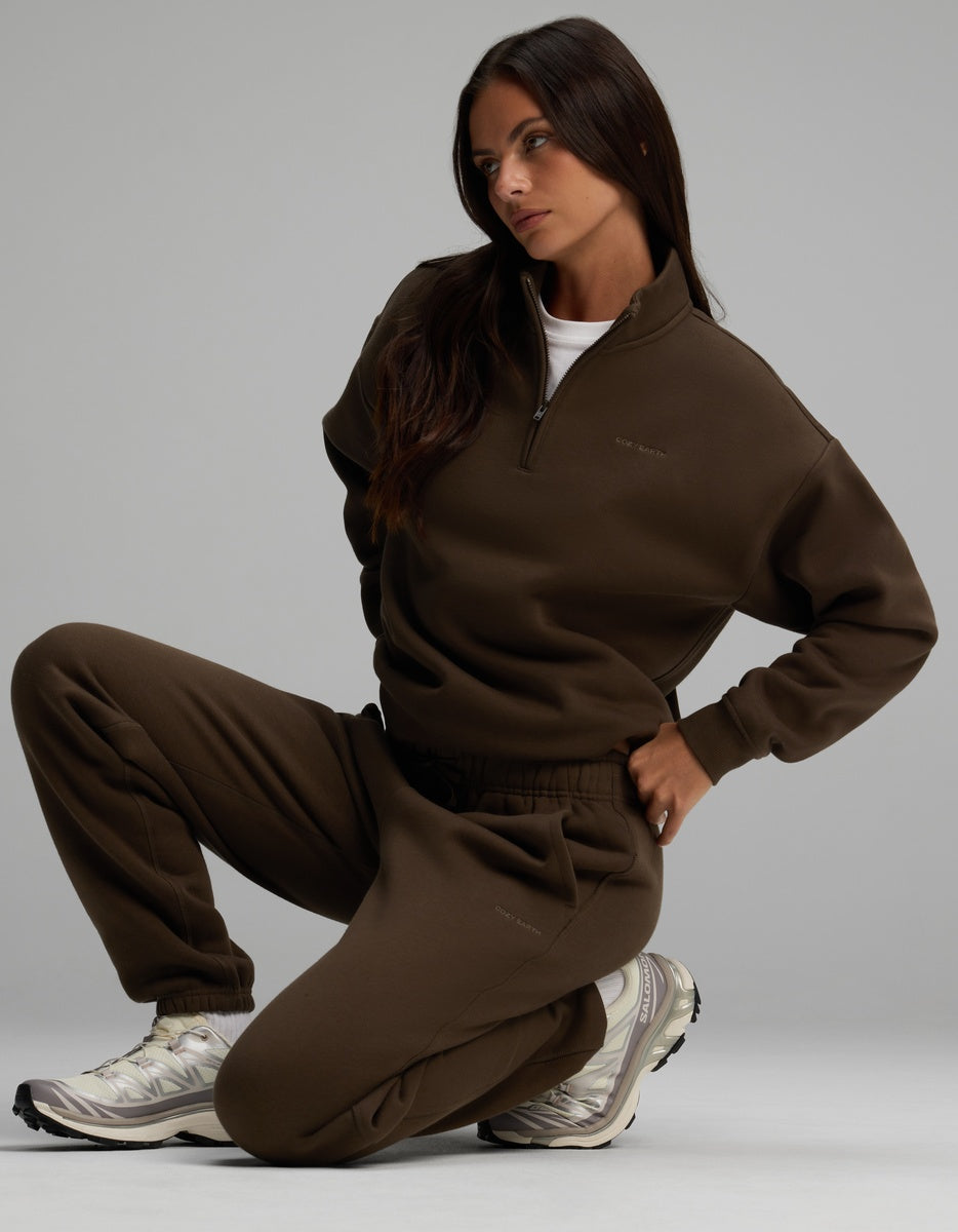 A person in a dark brown Cozy Earth Women’s Cityscape Quarter Zip & Sweatpant Set, crouched while wearing off-white athletic sneakers.