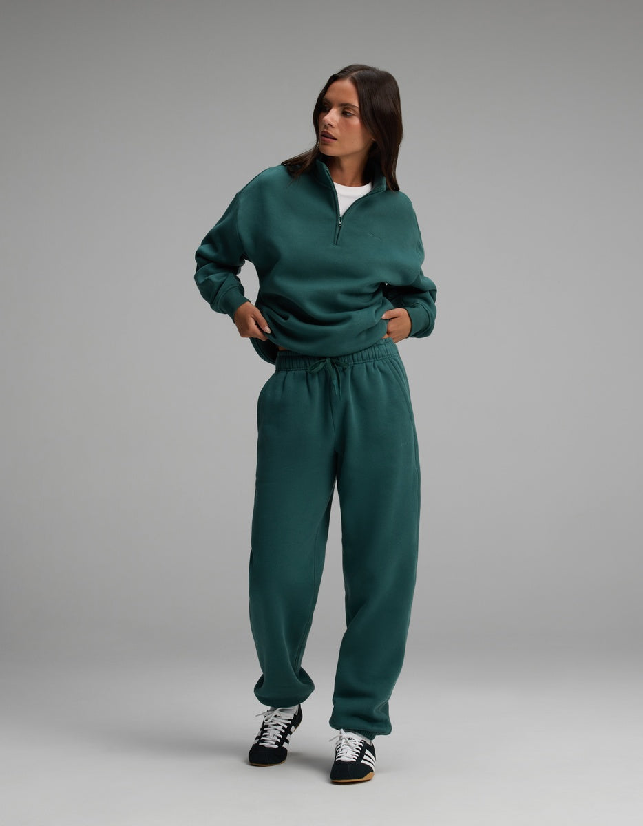 A person wearing a dark green Women’s Cityscape Quarter Zip & Sweatpant Set with black and white sneakers.