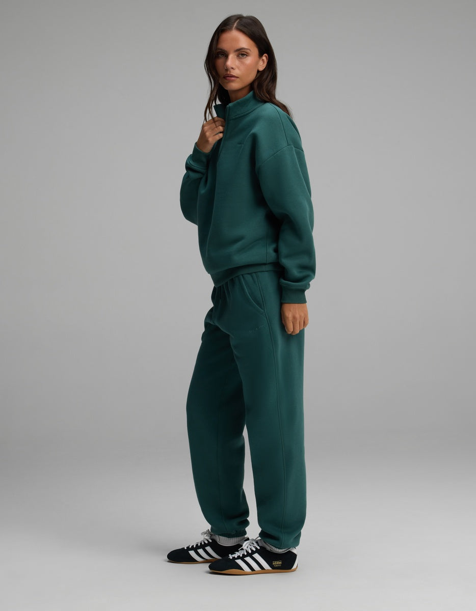 A woman wears a dark green Cozy Earth Women’s Cityscape Quarter Zip & Sweatpant Set with black and white sneakers.