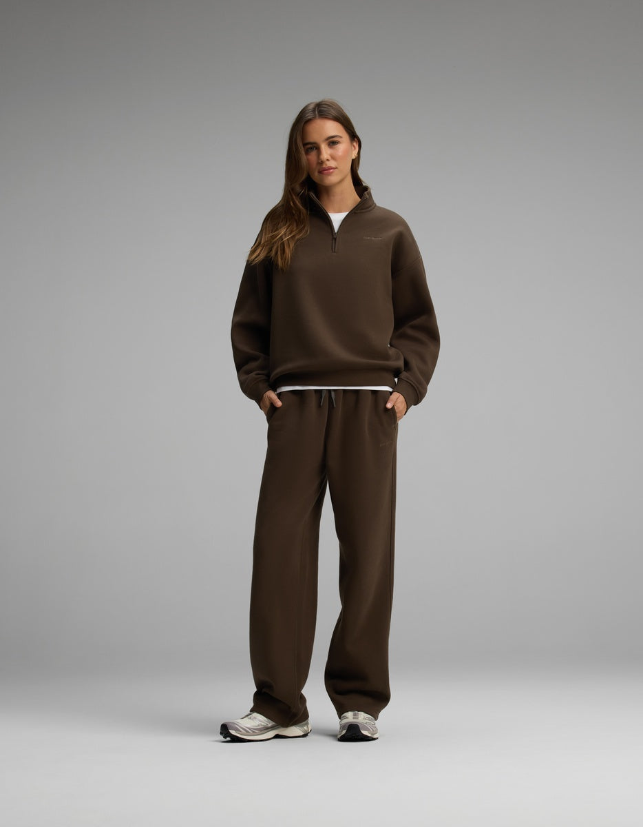 A woman wears a brown Cozy Earth Women’s Cityscape Quarter Zip & Wide Leg Pant Set, featuring a quarter-zip sweatshirt over a white t-shirt with matching wide-leg sweatpants and sneakers.