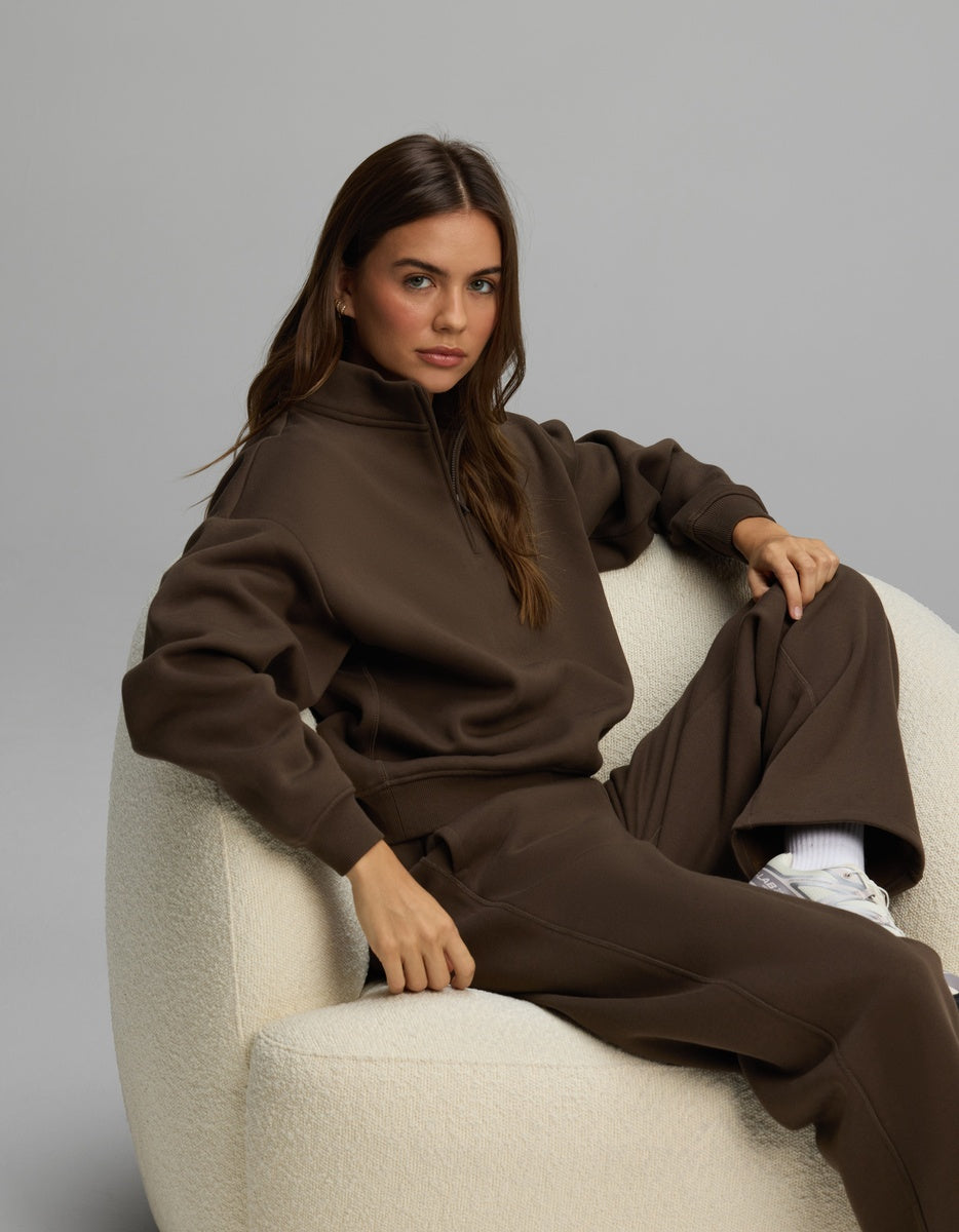 A woman wears a dark brown Cozy Earth Women’s Cityscape Quarter Zip & Wide Leg Pant Set while sitting in a white textured armchair.