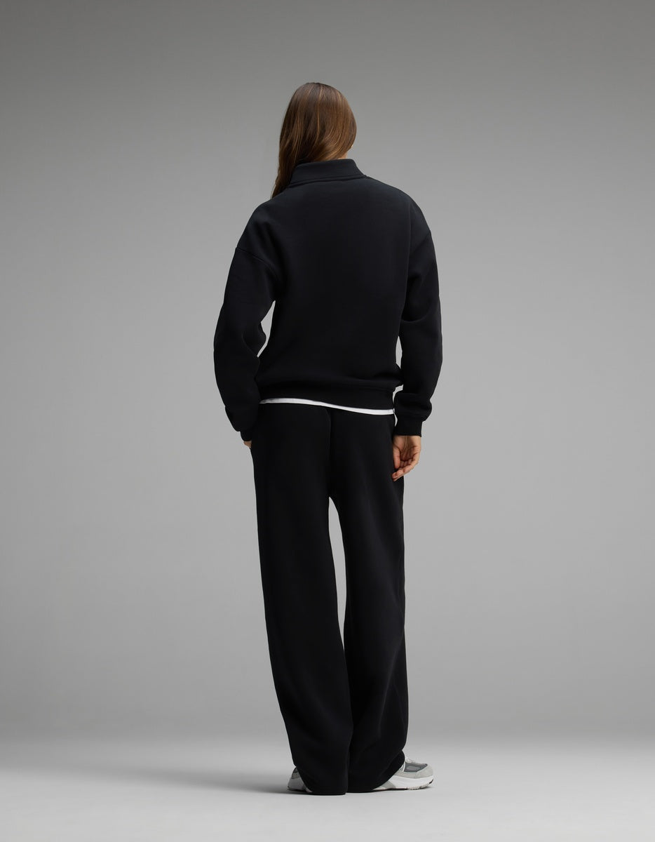 Seen from behind, a person wears a black Women’s Cityscape Quarter Zip & Wide Leg Pant Set consisting of a black quarter-zip sweatshirt over a white shirt, paired with matching black wide-leg pants.