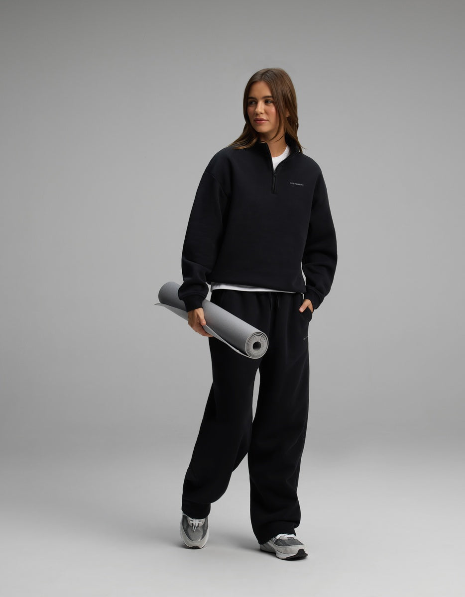 A woman wearing a black Cozy Earth Women’s Cityscape Quarter Zip & Wide Leg Pant Set, holding a rolled-up grey yoga mat.
