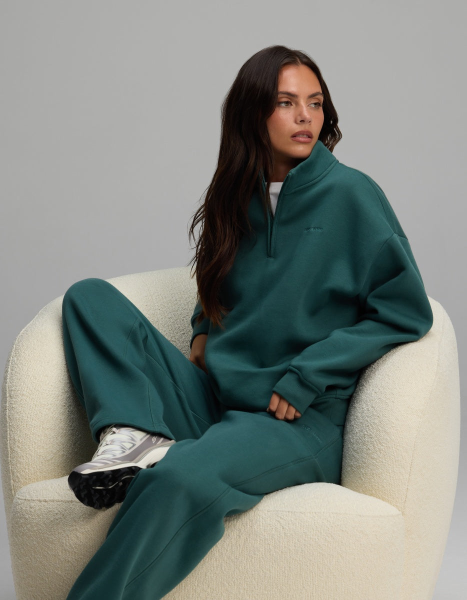 A woman wearing a deep green Cozy Earth Women’s Cityscape Quarter Zip & Wide Leg Pant Set sits in a white armchair.