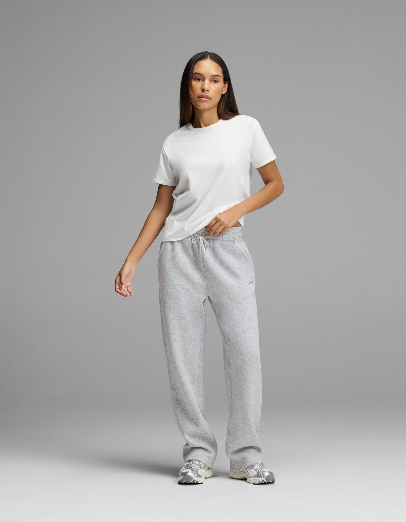 A person stands against a plain gray background wearing a white t-shirt, Cozy Earth Women’s Cityscape 2.0 Wide Leg Pant in light gray, and gray sneakers. They have long, straight hair and a neutral expression with one hand in their pocket.|Color:Heather Grey