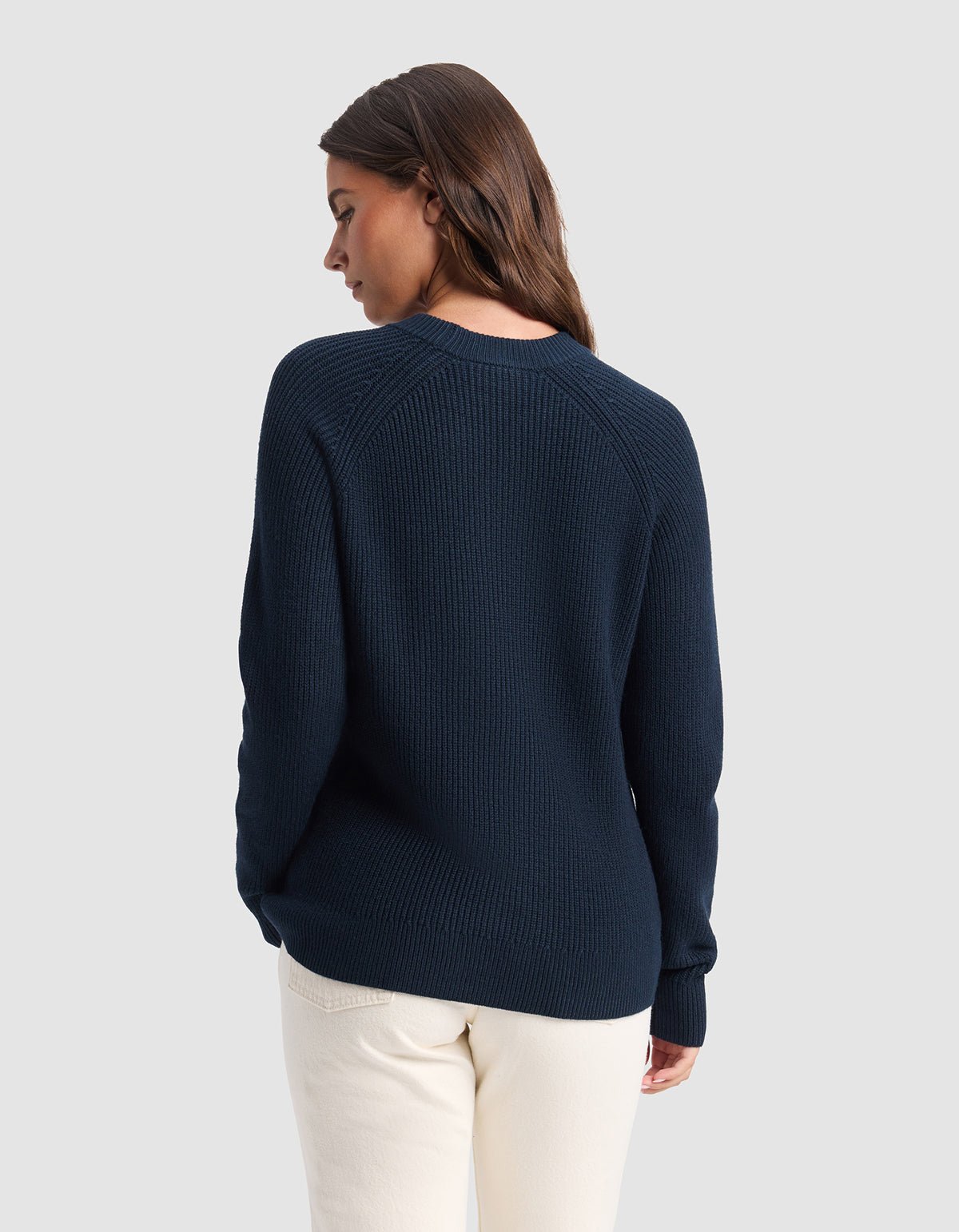 A woman with long brown hair stands with her back to the camera, wearing Cozy Earth's Women's Classic Crewneck in navy blue and off-white pants against a light grey background.|Color:Eclipse