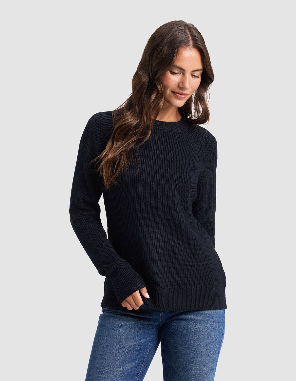 A woman with long brown hair models the Cozy Earth Women's Classic Crewneck, wearing blue jeans and standing against a light gray background, looking down with a slight smile and one hand in her pocket. |Color:Jet Black
