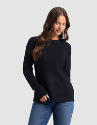 A woman with long brown hair models the Cozy Earth Women's Classic Crewneck, wearing blue jeans and standing against a light gray background, looking down with a slight smile and one hand in her pocket. |Color:Jet Black