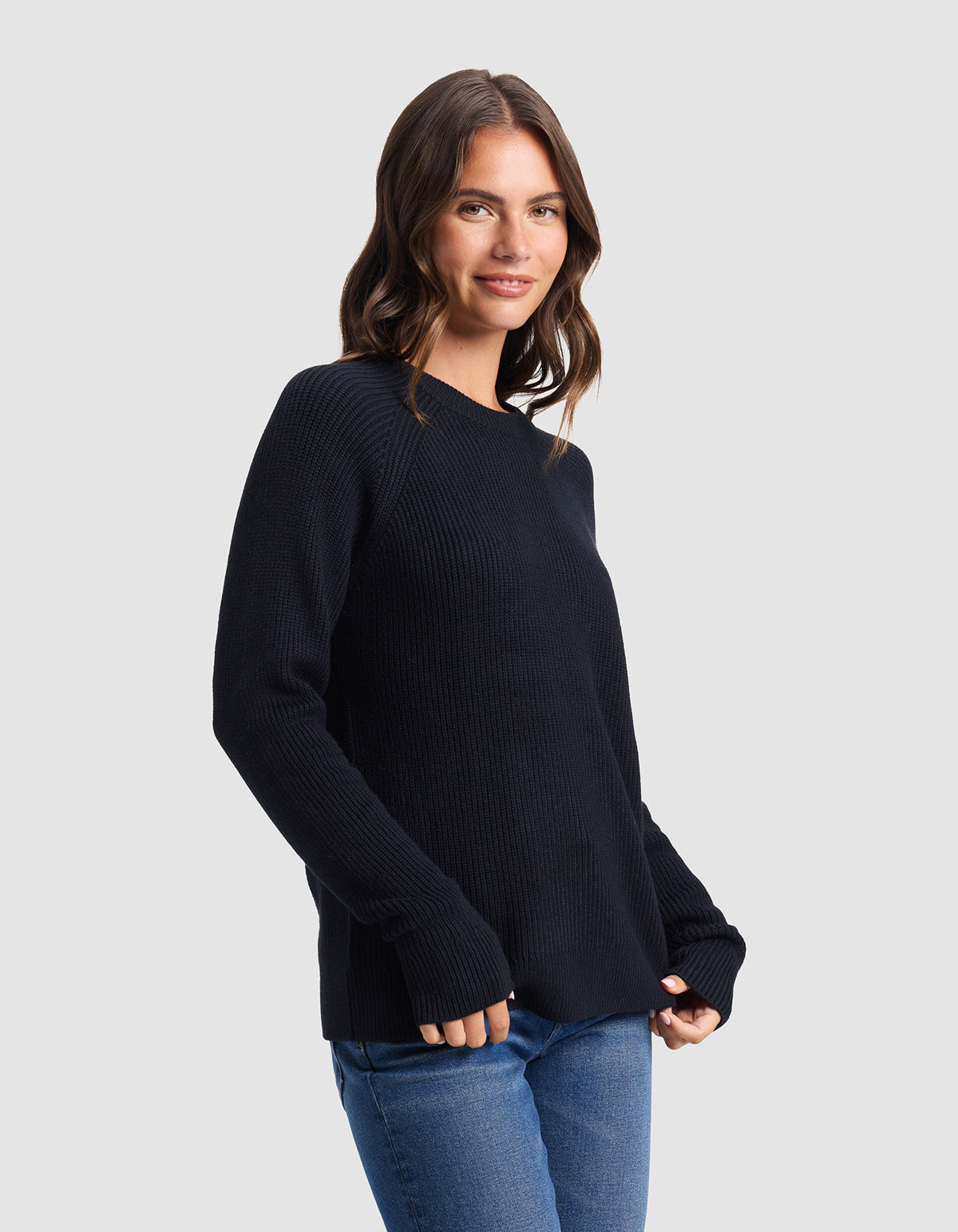 A woman with wavy brown hair smiles softly while wearing the Cozy Earth Women's Classic Crewneck in black and blue jeans, standing against a plain light gray background with her hands relaxed by her sides.|Color:Jet Black
