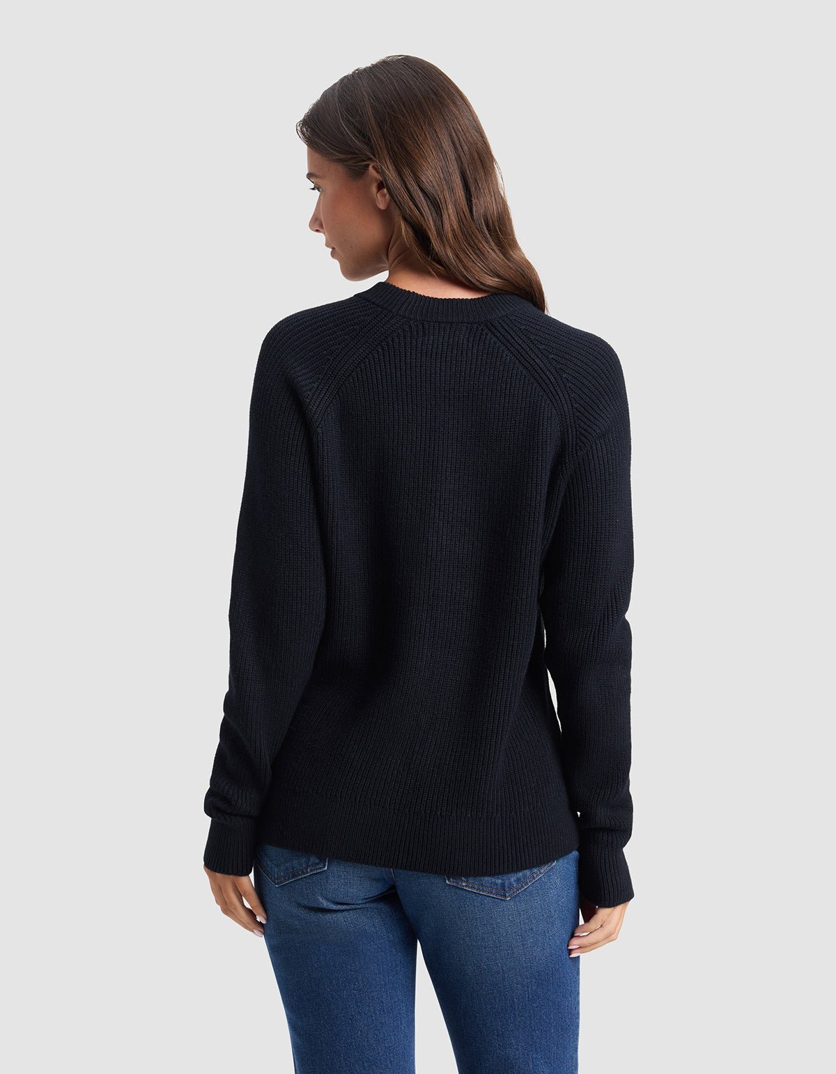 A woman with long brown hair, wearing the Cozy Earth Women's Classic Crewneck and blue jeans, stands facing away from the camera against a plain light gray background.