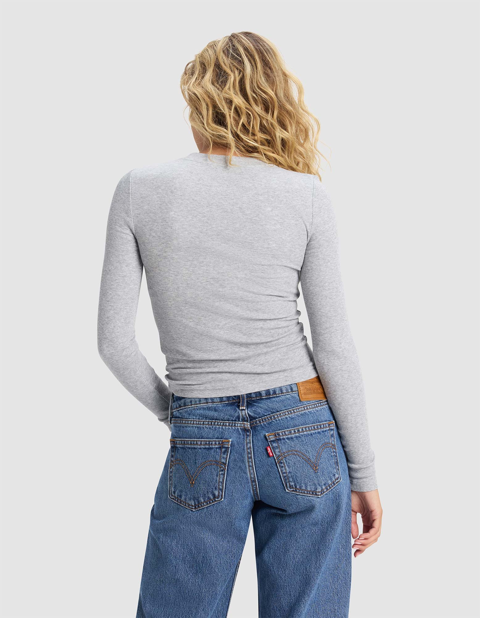 A woman with wavy blonde hair, wearing Cozy Earth's Women's Fine Ribbed Long Sleeve Tee in light gray and blue jeans, stands with her back to the camera against a plain light background.