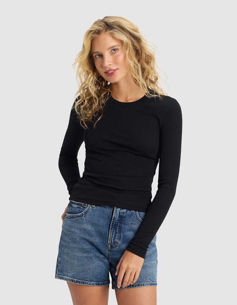 A woman with wavy blonde hair models the Cozy Earth Women's Fine Ribbed Long Sleeve Tee in black, paired with blue denim shorts, standing with one hand in her pocket against a plain light gray background.