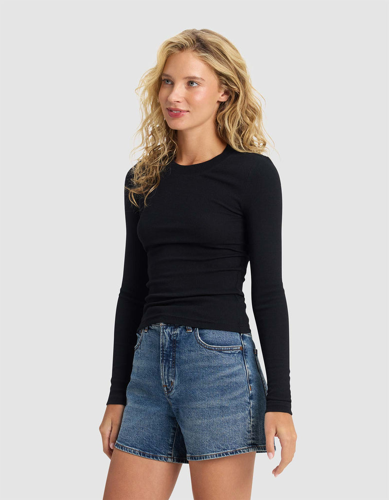 A woman with wavy blonde hair wears the Cozy Earth Women's Fine Ribbed Long Sleeve Tee in black and denim shorts, standing against a plain light gray background. She faces slightly to the side with a soft smile.