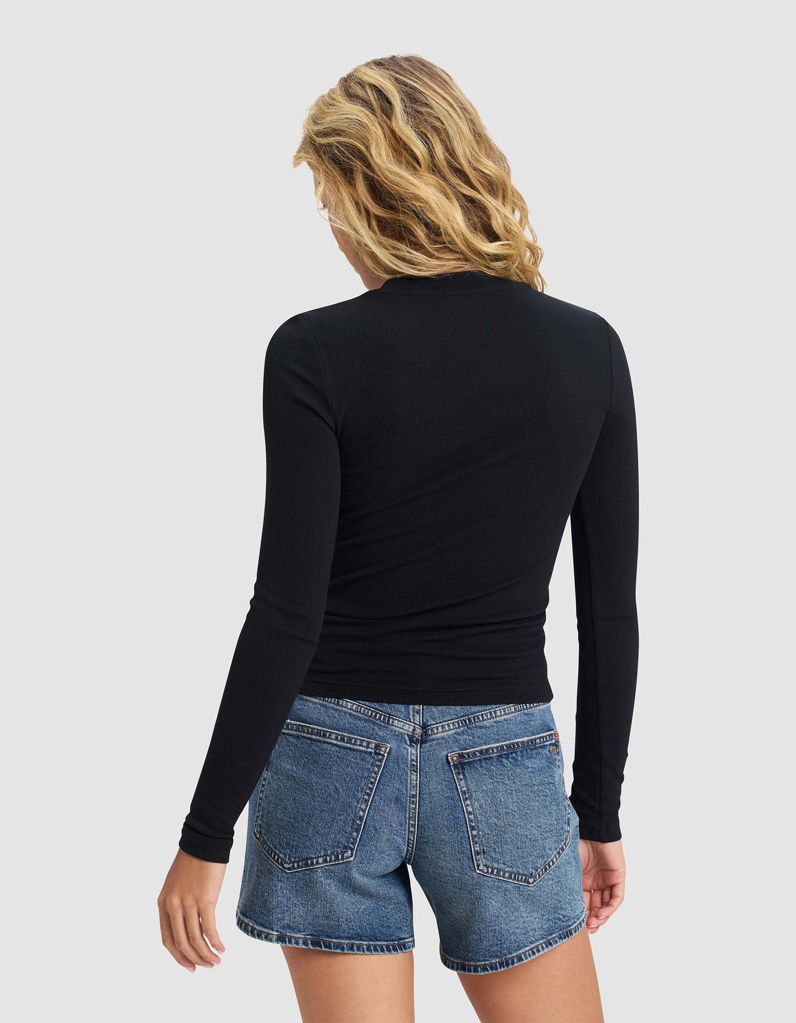A woman with wavy blonde hair is seen from behind, wearing the Cozy Earth Women's Fine Ribbed Long Sleeve Tee in black and blue denim shorts, standing against a plain light gray background.|Color:Jet Black