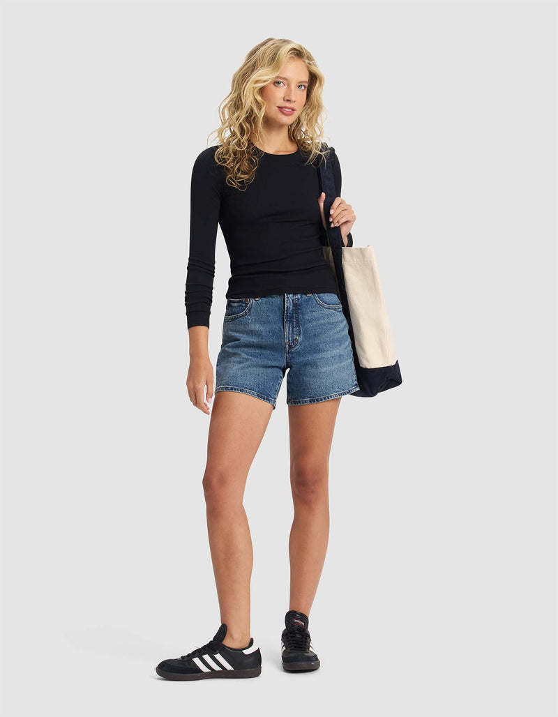 A woman with wavy blonde hair wears the Cozy Earth Women's Fine Ribbed Long Sleeve Tee in black, denim shorts, black Adidas sneakers, and carries a beige tote bag. She stands against a plain light gray background.