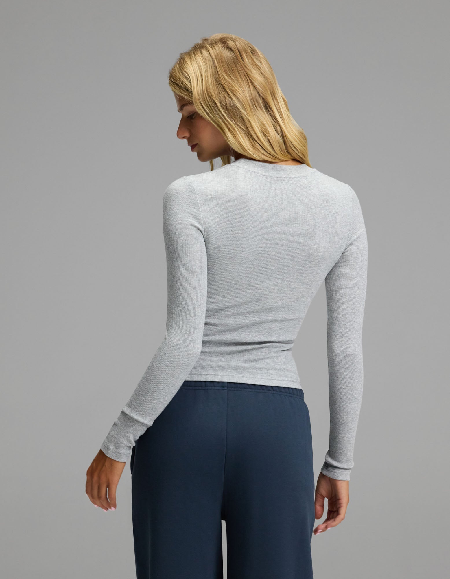 A woman with long blonde hair, wearing the Cozy Earth Women’s Fine Ribbed Long Sleeve Tee in light gray and dark blue pants, stands facing away and looks over her shoulder against a plain gray background.