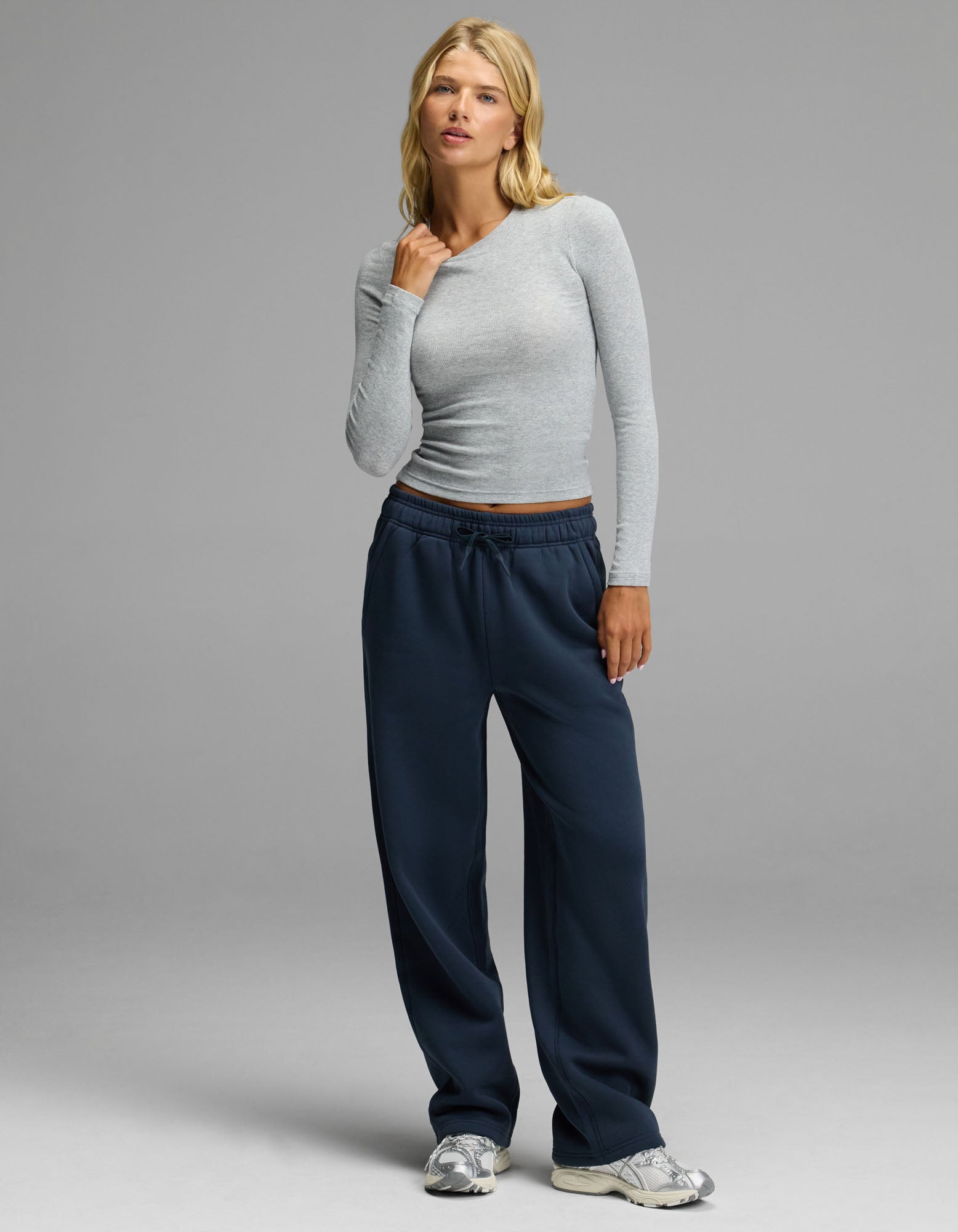 A woman with blonde hair and a neutral expression stands against a gray background, wearing the Cozy Earth Women’s Fine Ribbed Long Sleeve Tee in light gray, paired with loose navy sweatpants and silver sneakers.