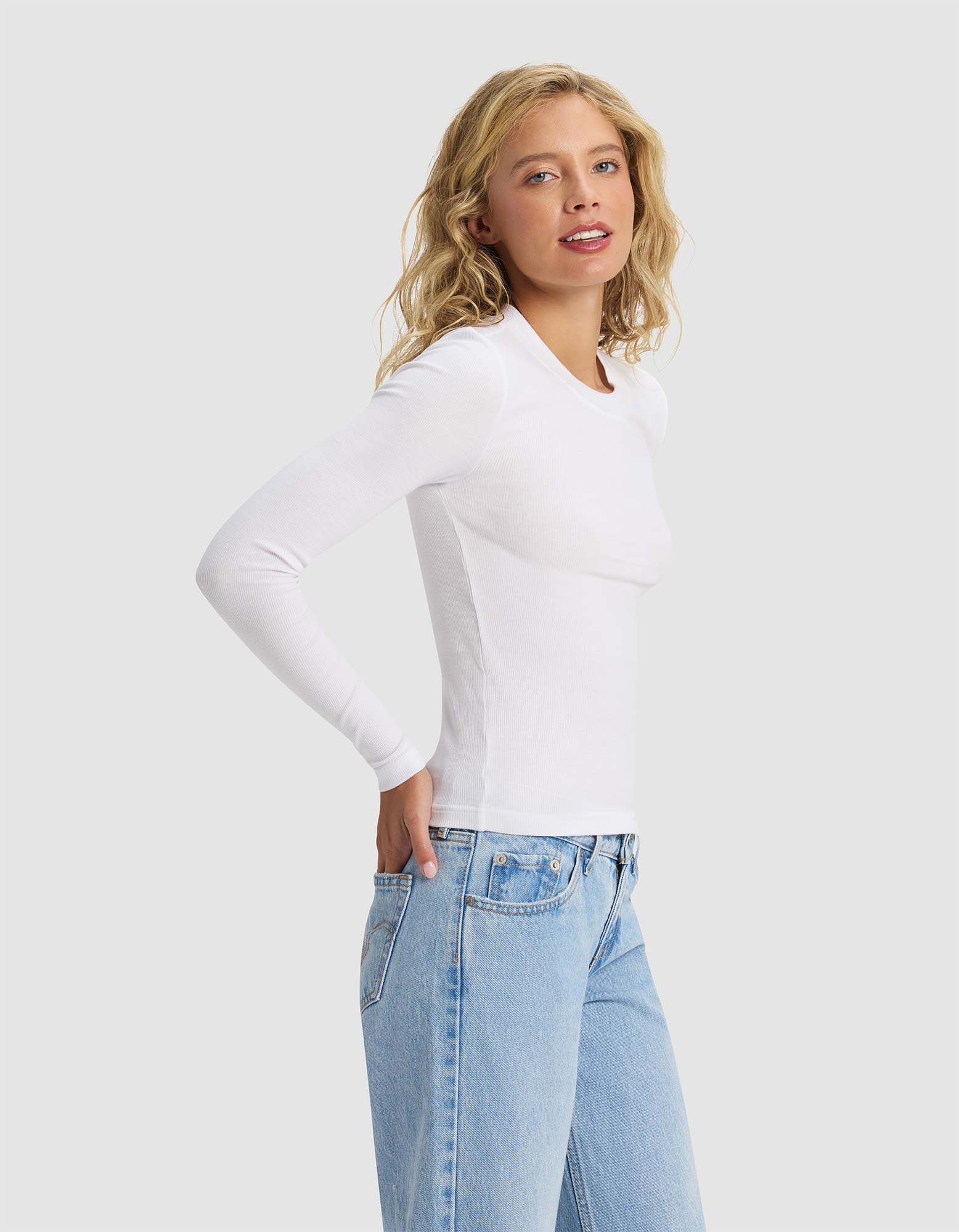 A woman with wavy blonde hair models the Cozy Earth Women's Fine Ribbed Long Sleeve Tee in white and light blue jeans, standing against a plain light gray background and looking slightly to the side.