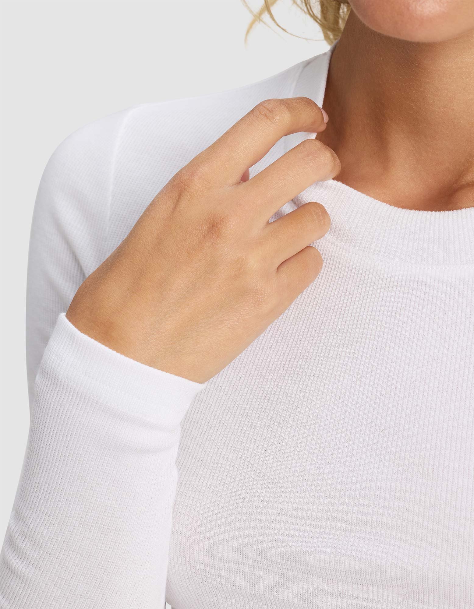 A close-up shows a person in the Cozy Earth Women's Fine Ribbed Long Sleeve Tee in white, gently touching the neckline. Their face is partly out of frame against a light gray background.