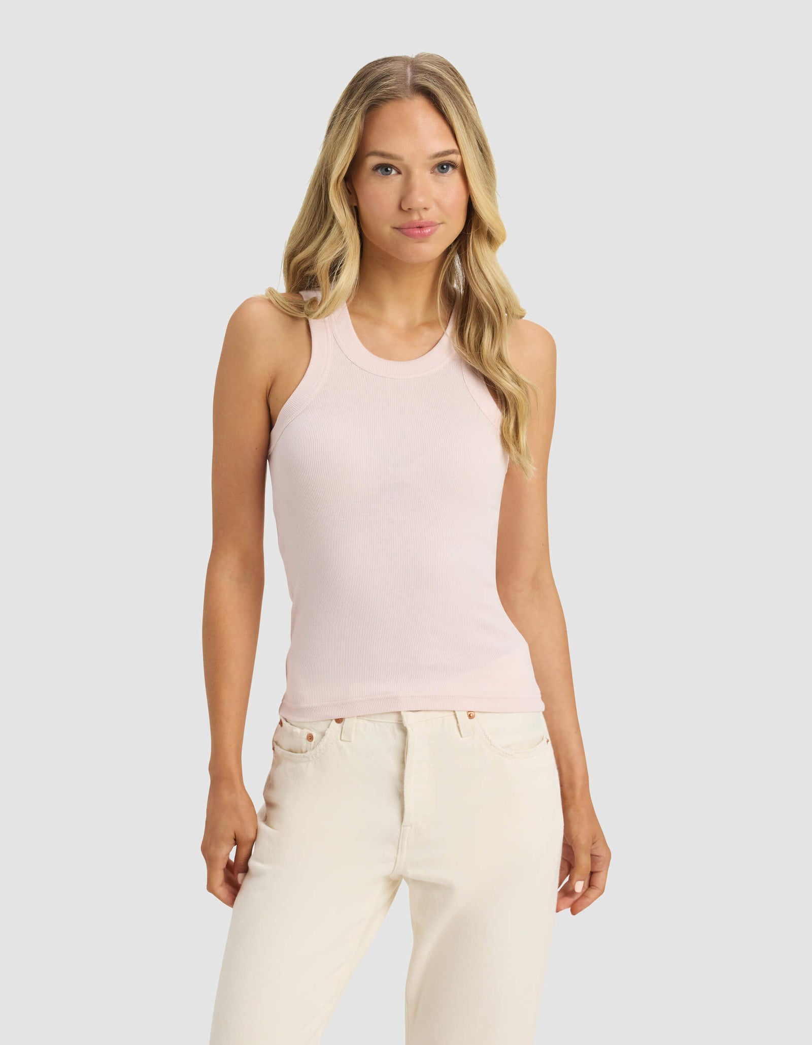 A woman with long blonde hair wears Cozy Earth's Women's Fine Ribbed Tank in light pink and off-white jeans, standing against a plain light gray background and facing forward with a neutral expression.