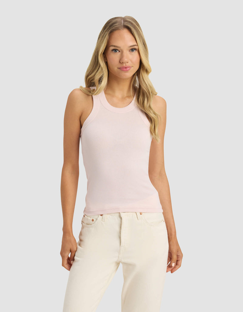 A woman with long blonde hair wears Cozy Earth's Women's Fine Ribbed Tank in light pink and off-white jeans, standing against a plain light gray background and facing forward with a neutral expression.