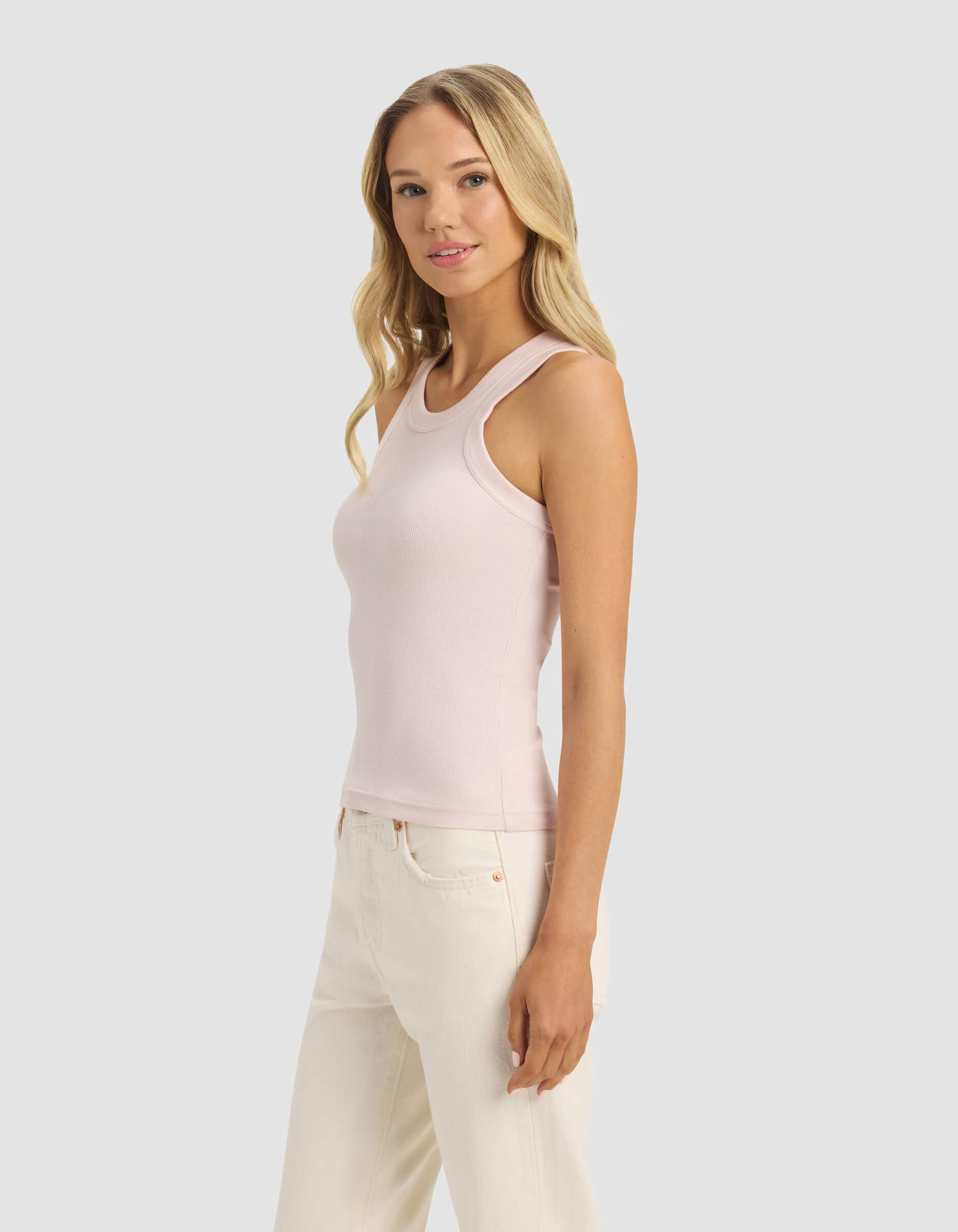 A woman with long blonde hair wears the Cozy Earth Women's Fine Ribbed Tank in light pink with off-white pants, standing against a plain light gray background and smiling softly.