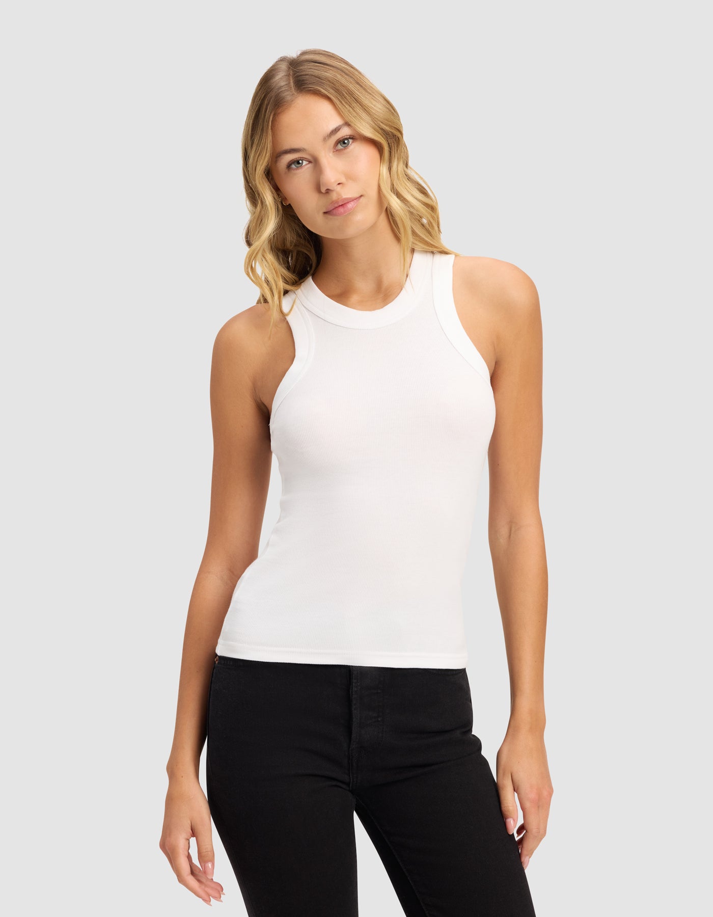 A woman with wavy blonde hair wears the Cozy Earth Women's Fine Ribbed Tank in white with black pants, standing against a plain light gray background and looking forward with a neutral expression.|Color:White