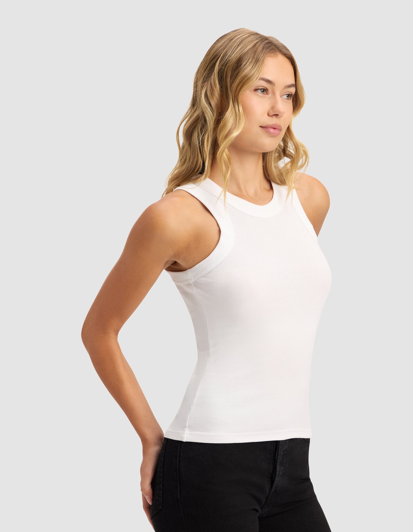 A woman with wavy blonde hair wears the Cozy Earth Women's Fine Ribbed Tank in white and black pants, standing against a plain light gray background, looking slightly to the side with her hands behind her back.