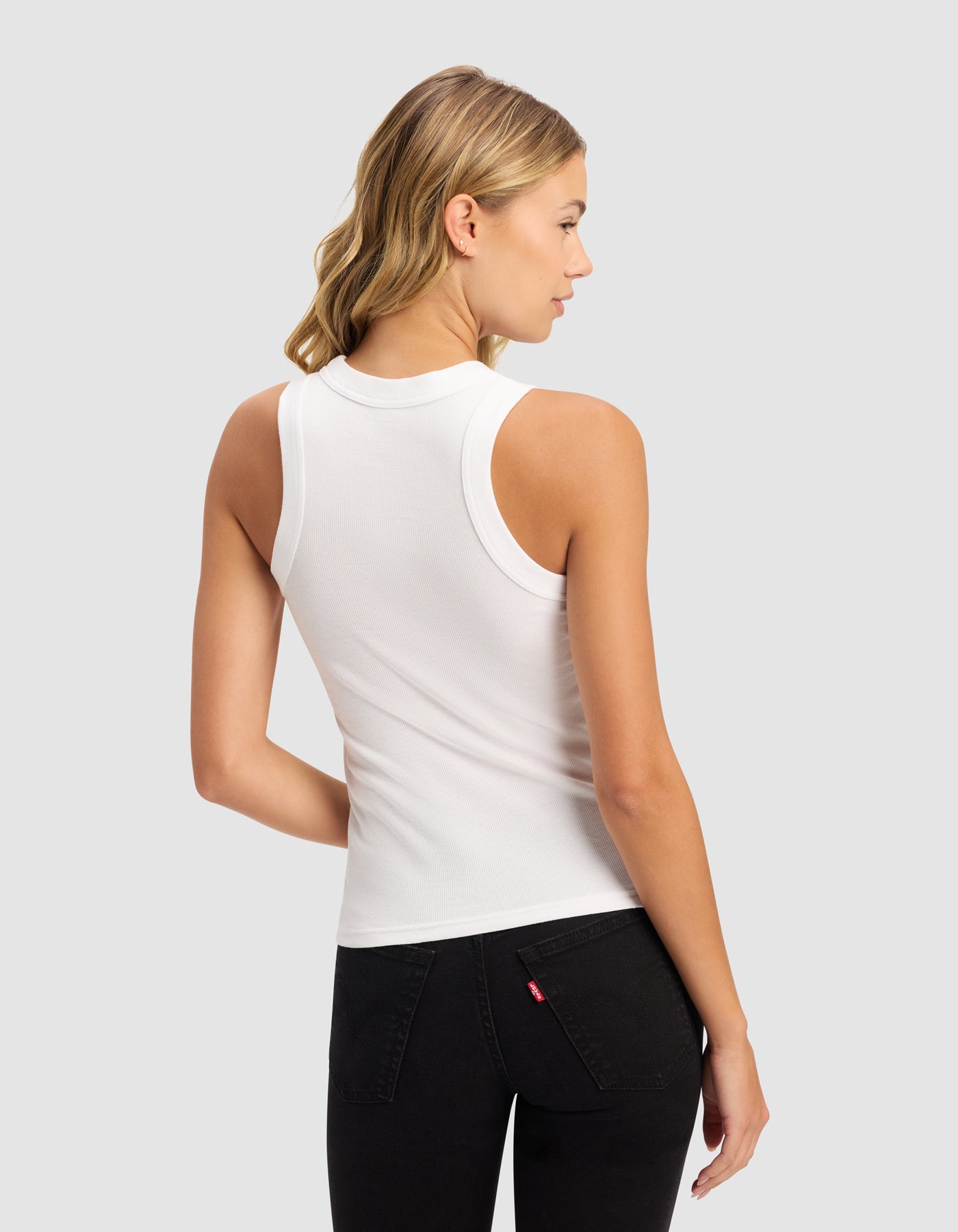 A woman with blonde wavy hair stands with her back to the camera, wearing Cozy Earth's Women's Fine Ribbed Tank in white and black jeans, set against a light gray background.|Color:White