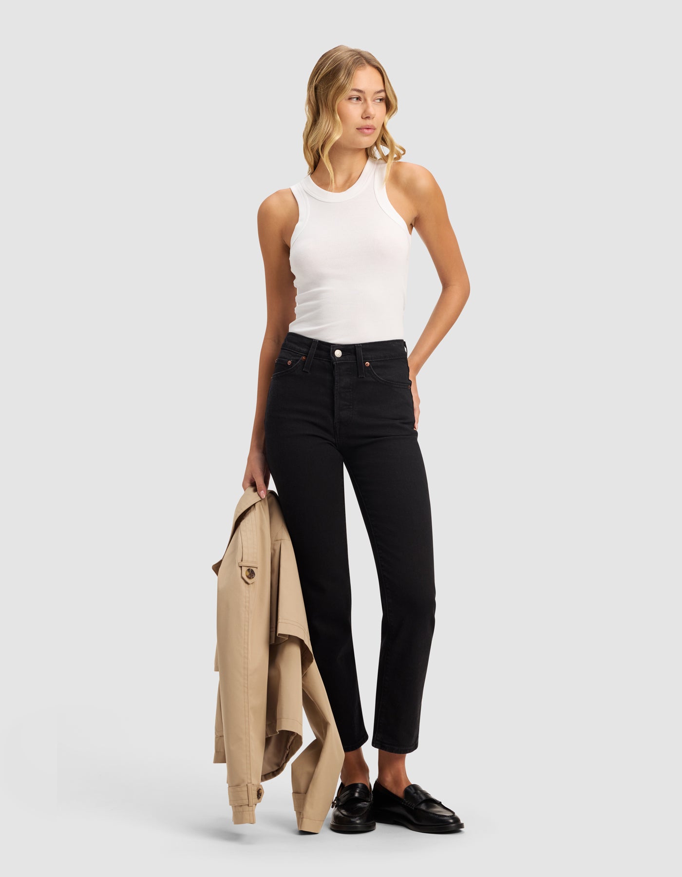 A woman stands against a plain background, wearing the Cozy Earth Women's Fine Ribbed Tank in white, paired with high-waisted black jeans and black loafers. She holds a beige trench coat in her left hand and gazes slightly to the side.|Color:White