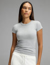A woman with long brown hair wears the Cozy Earth Women’s Fine Ribbed Tee in light grey and white drawstring pants, standing against a plain grey background and looking slightly to her left with a neutral expression.|Color:Heather Grey