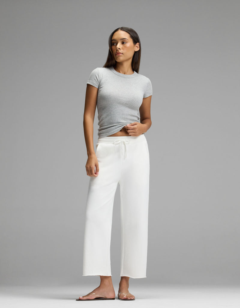A woman stands against a plain gray background, wearing the Cozy Earth Women’s Fine Ribbed Tee in gray, paired with loose white pants and sandals. She looks slightly to the side with a neutral expression.