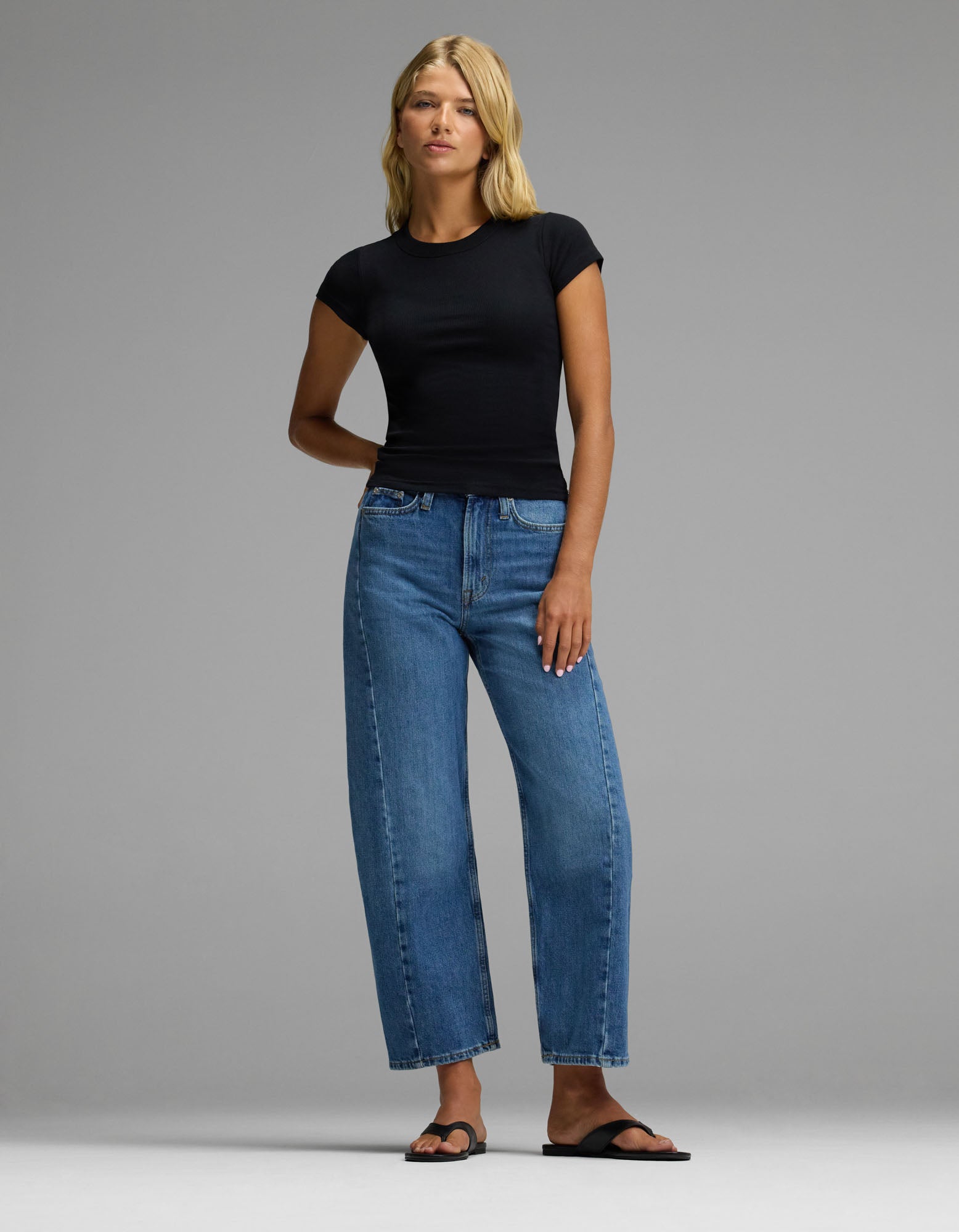 A woman with blonde hair stands against a plain background wearing the Cozy Earth Women’s Fine Ribbed Tee in black, paired with high-waisted blue jeans and black sandals. Her left arm rests behind her back.