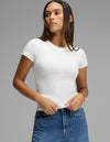 A woman wearing the Cozy Earth Women’s Fine Ribbed Tee in white and blue jeans stands against a gray background, looking to the side with her hands holding the hem of her shirt. She has long, straight hair and accessorizes with a gold watch.|Color:White