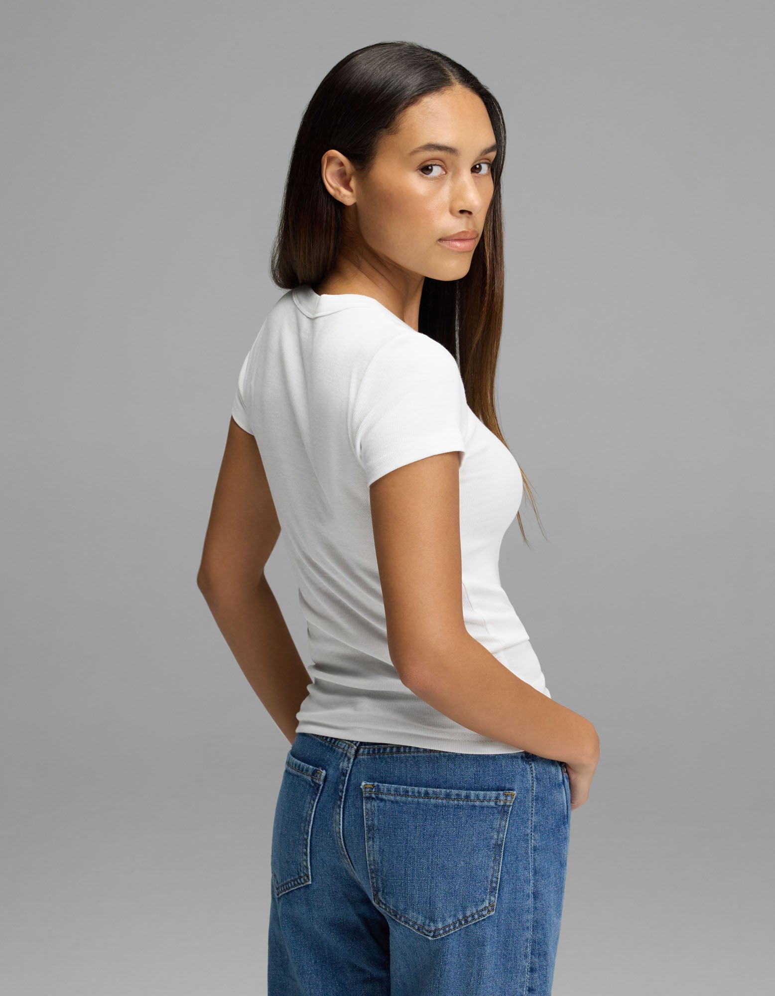 A woman with long brown hair wears the Cozy Earth Women’s Fine Ribbed Tee in white and blue jeans, standing sideways and looking over her shoulder against a plain gray background.