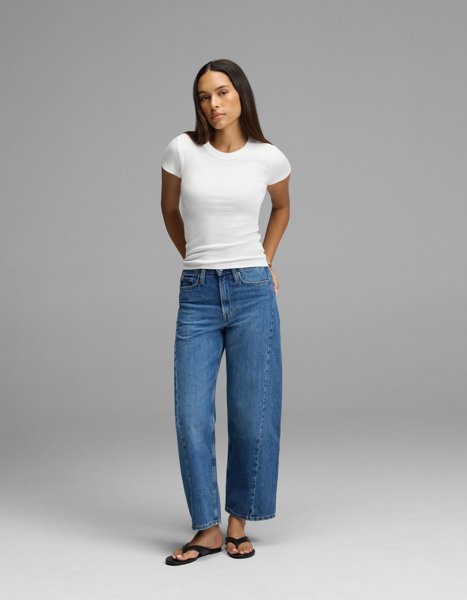 A woman stands against a plain gray background wearing the Cozy Earth Women’s Fine Ribbed Tee, styled with blue wide-leg jeans and black sandals. Her arms are behind her back, and she has long straight hair.