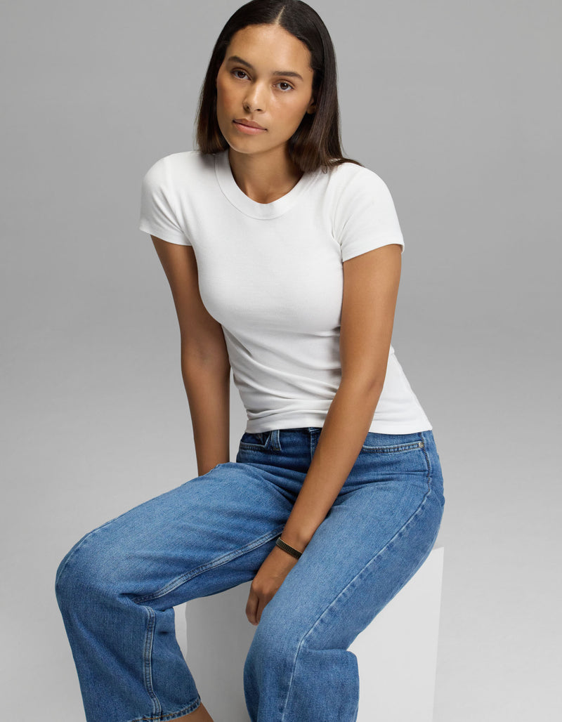 A woman with straight dark hair, dressed in the Cozy Earth Women’s Fine Ribbed Tee and blue jeans, sits on a white block against a plain gray background, calmly looking at the camera.