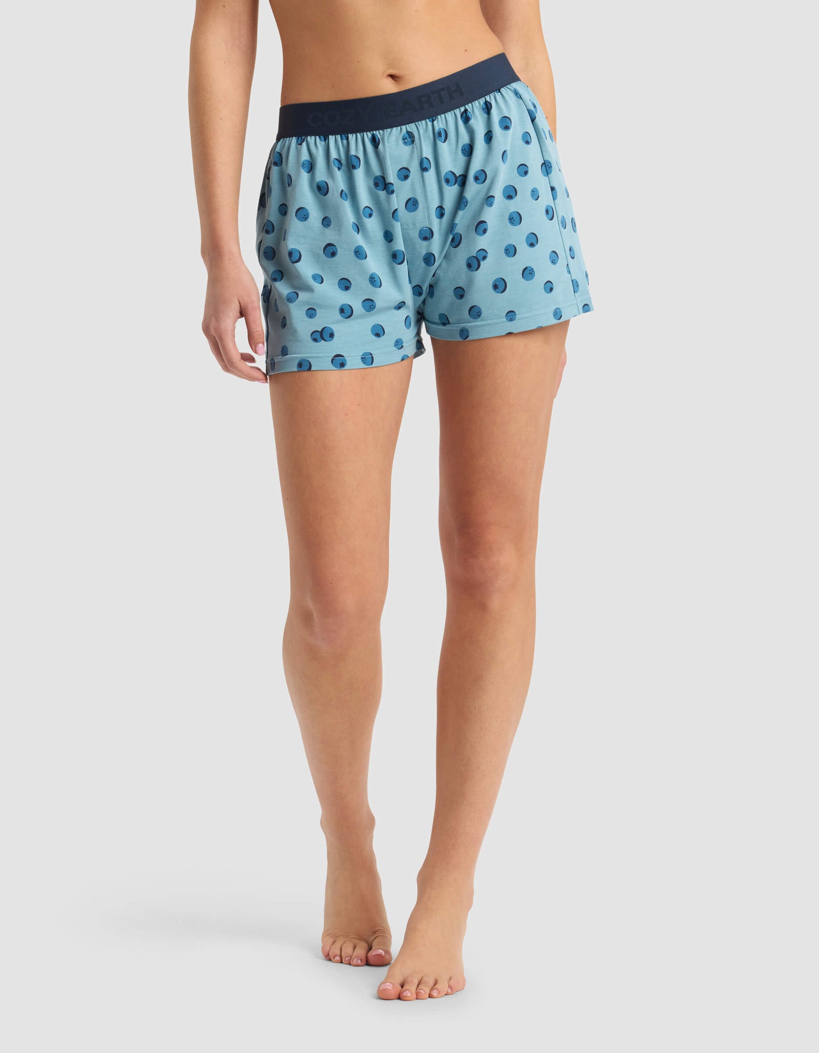 A person stands barefoot against a white background, wearing Cozy Earth's Women's First Layer Pajama Boxer Short in light blue with a dark blue polka dot pattern and a dark blue waistband.