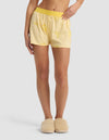 A person wearing Cozy Earth's Women's First Layer Pajama Boxer Short in yellow with lemon slice prints, a matching waistband, and beige fuzzy slippers stands against a plain light background. Only the lower torso and legs are visible.|Color:Lemons