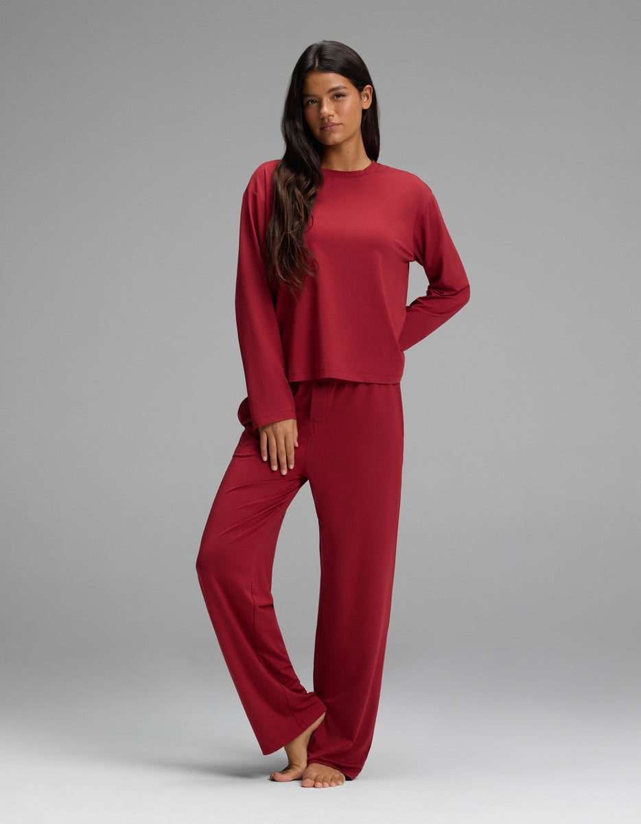 A woman wears a dark red Women’s First Layer Long Sleeve Sleep Tee & Pajama Pant Set featuring a long-sleeve, crewneck pajama top and matching full-length pajama pants.