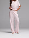 A woman stands barefoot wearing the Cozy Earth Women’s First Layer Pajama Pant in Ballet, a loose-fitting, light pink short-sleeve pajama set with wide-leg pants. She holds up part of her shirt, and her head is not visible in the image.|Color:Ballet