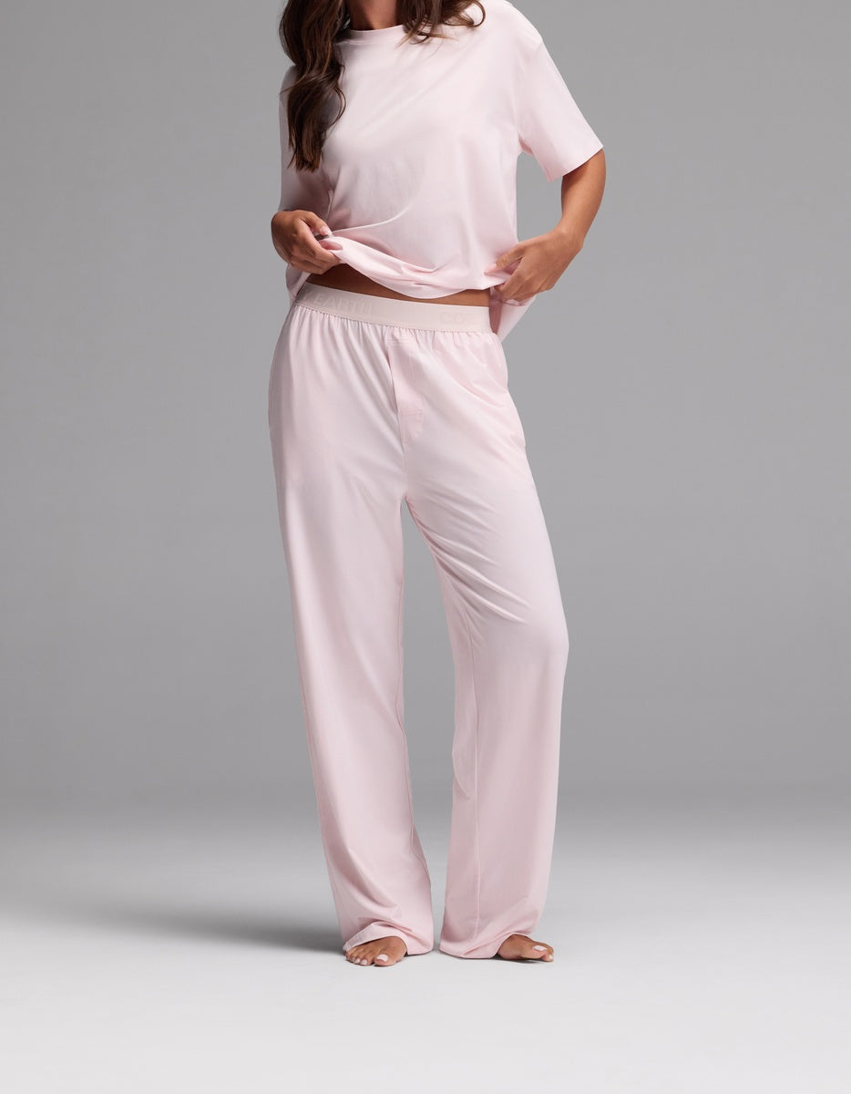 A woman stands barefoot wearing the Cozy Earth Women’s First Layer Pajama Pant in Ballet, a loose-fitting, light pink short-sleeve pajama set with wide-leg pants. She holds up part of her shirt, and her head is not visible in the image.|Color:Ballet