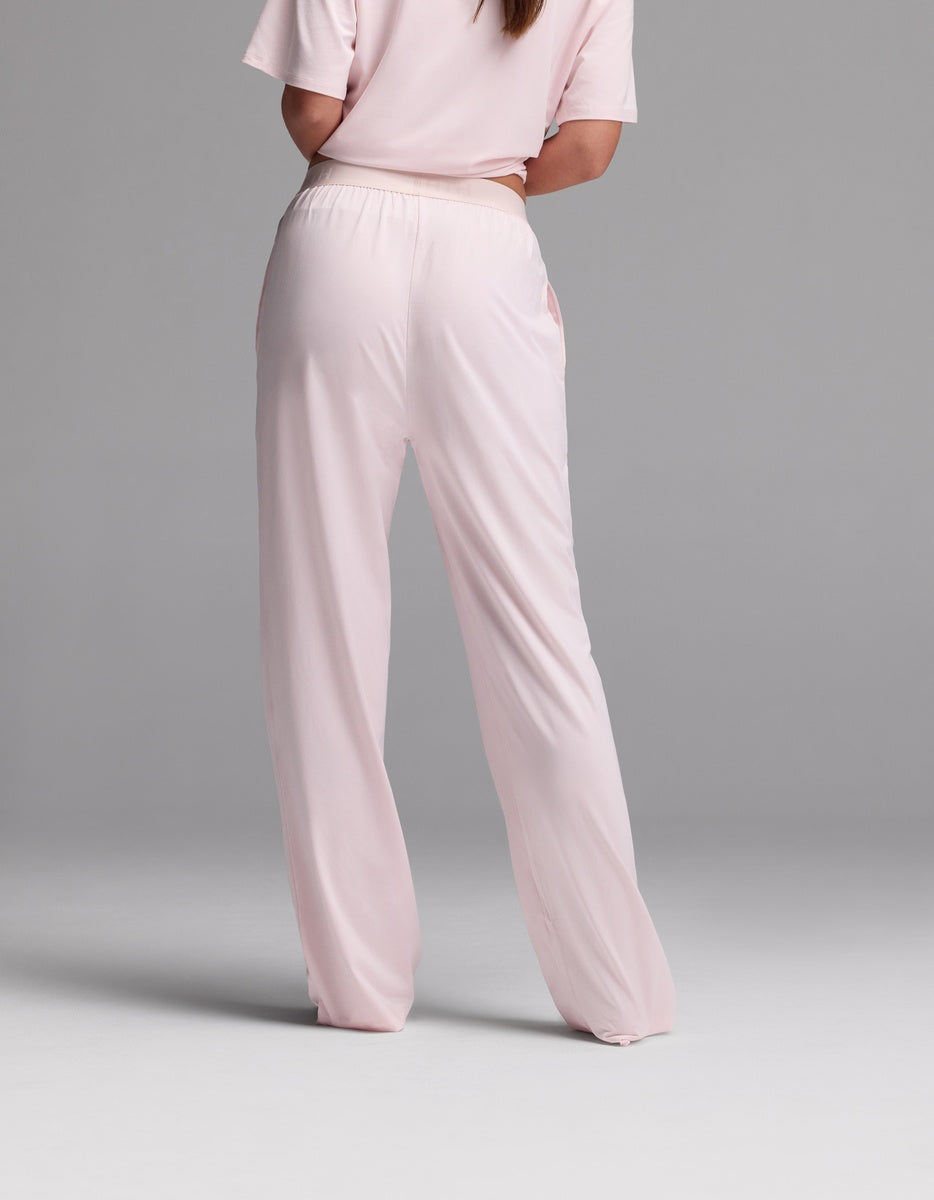 A woman is shown from the back, standing against a plain gray background, wearing the Cozy Earth Women’s First Layer Pajama Pant in light pink with a matching short-sleeve top.|Color:Ballet