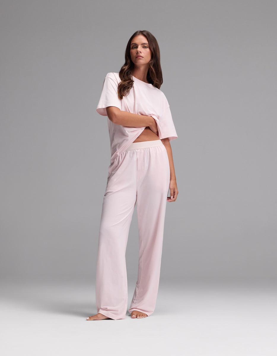 A woman stands barefoot against a gray background, wearing the Cozy Earth Women’s First Layer Pajama Pant in light pink, paired with a loose-fitting, short-sleeved top. Her hair is styled in loose waves and her expression is neutral.