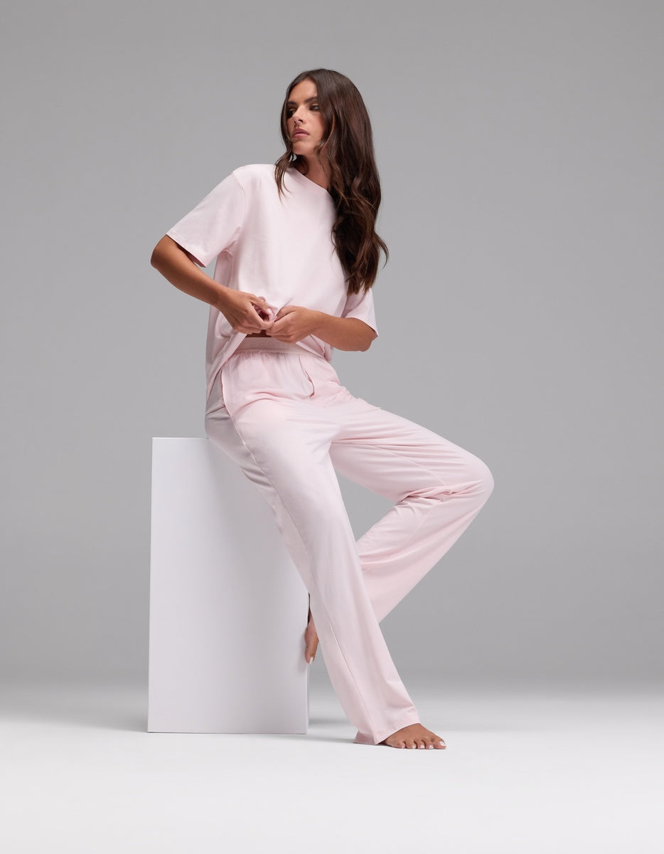 A woman with long brown hair wears the Cozy Earth Women’s First Layer Pajama Pant in light pink and sits barefoot on a white block against a plain gray background.