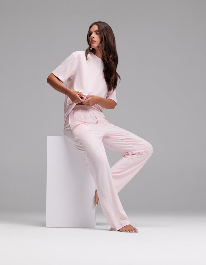 A woman with long brown hair wears the Cozy Earth Women’s First Layer Pajama Pant in light pink and sits barefoot on a white block against a plain gray background.