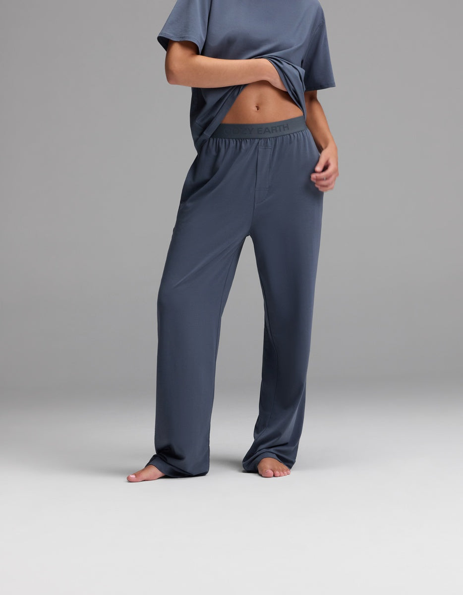 A person stands barefoot on a plain gray background, wearing Cozy Earth Women’s First Layer Pajama Pant in Dawn with a matching T-shirt, holding up part of the shirt to reveal the pajama pant’s waistband.