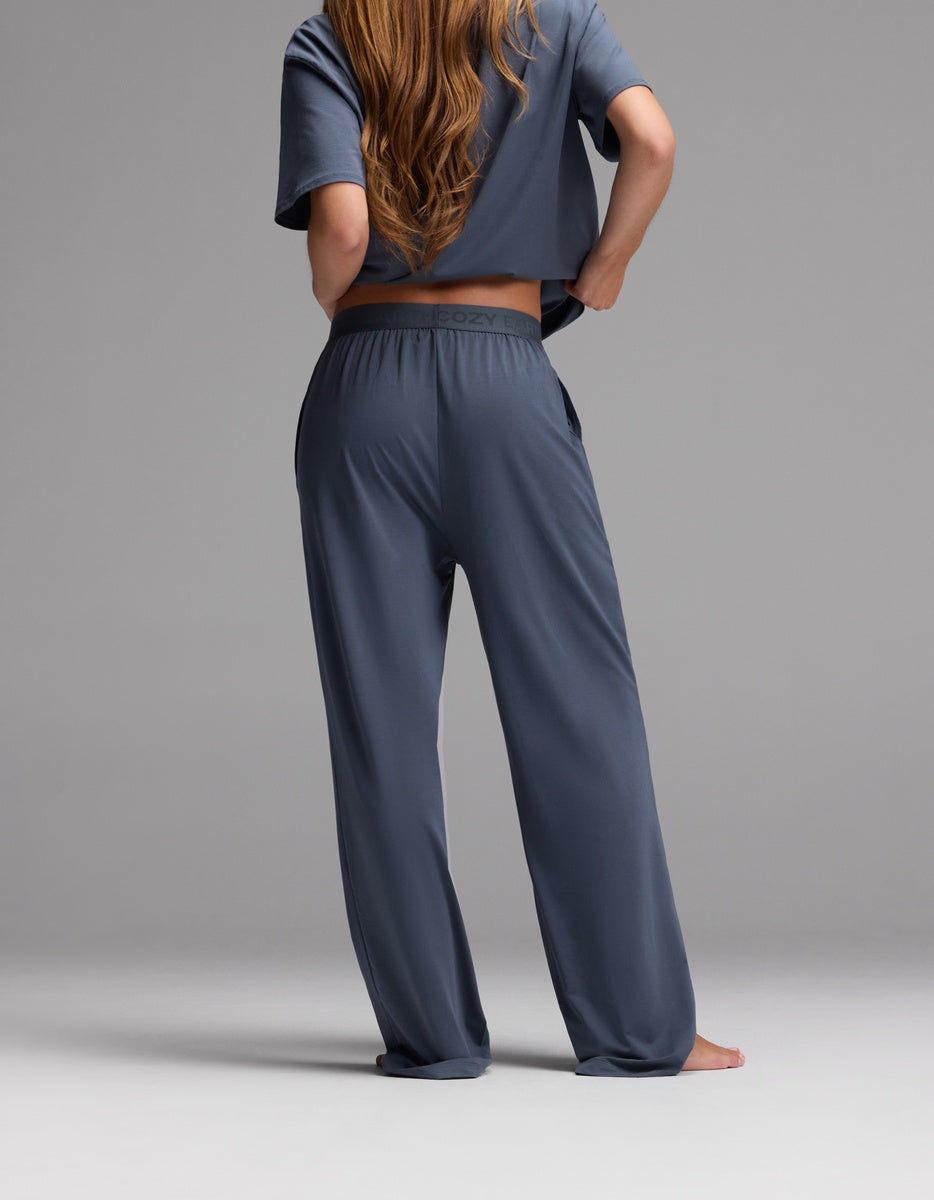 A woman with long hair is standing barefoot and facing away against a plain gray background while wearing the Cozy Earth Women’s First Layer Pajama Pant in slate blue, paired with a matching loose, short-sleeve top.