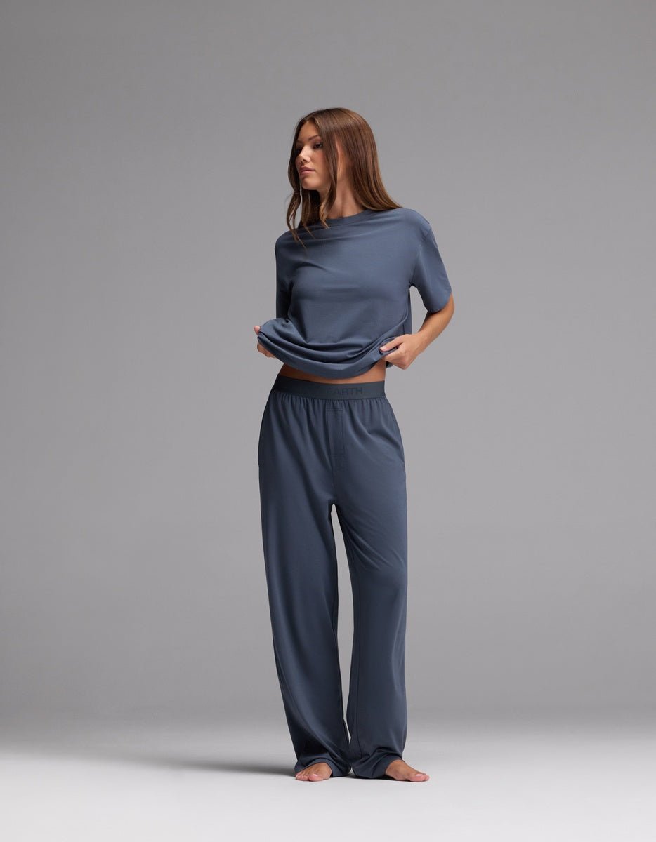A woman stands barefoot against a gray background, wearing Cozy Earth’s Women’s First Layer Pajama Pant in blue. She lifts her shirt slightly with one hand and gazes to the side with a neutral expression.
