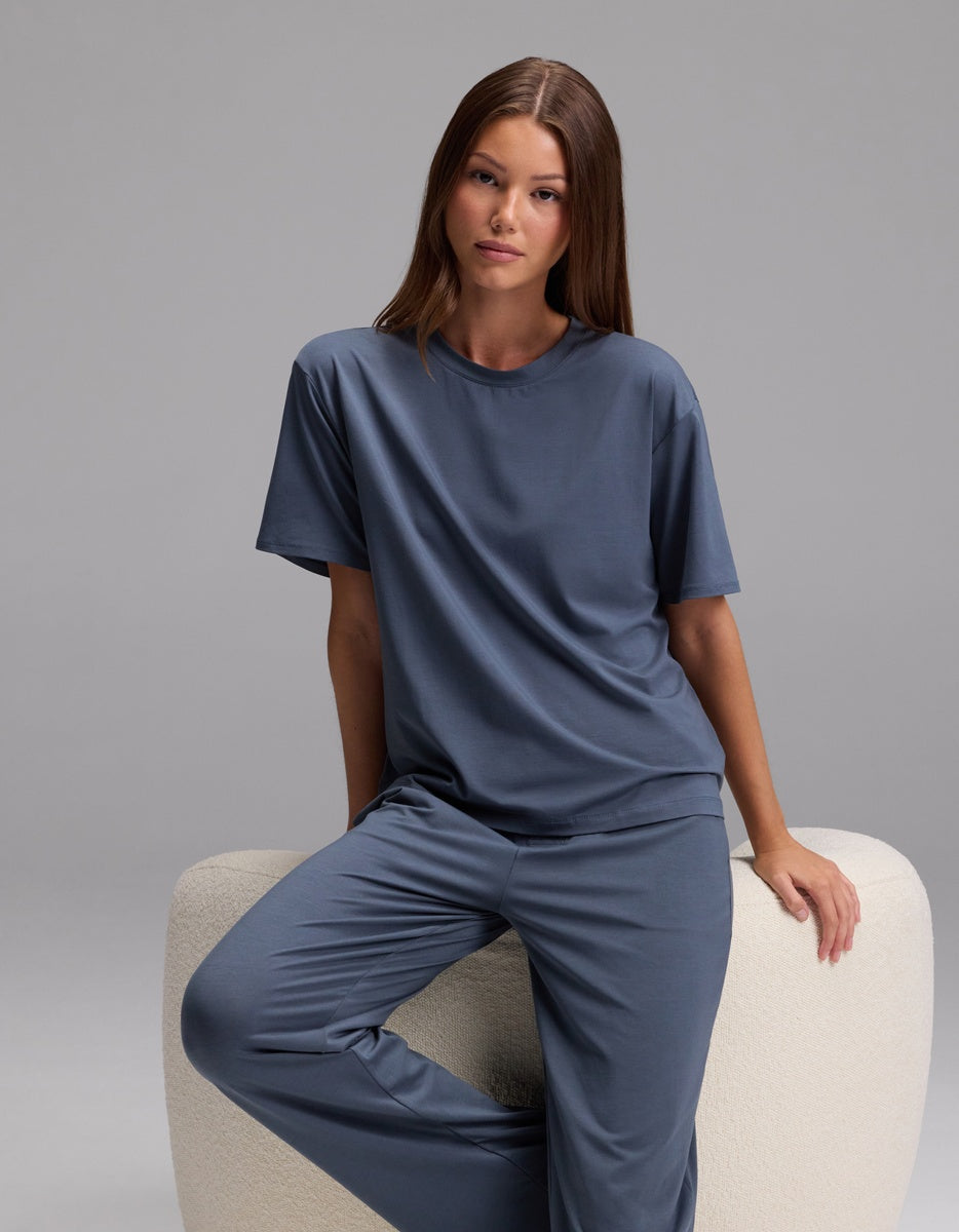 A woman with long brown hair wears Cozy Earth’s Women’s First Layer Pajama Pant and a matching loose blue top as she sits on a light-colored, curved chair against a plain gray background.